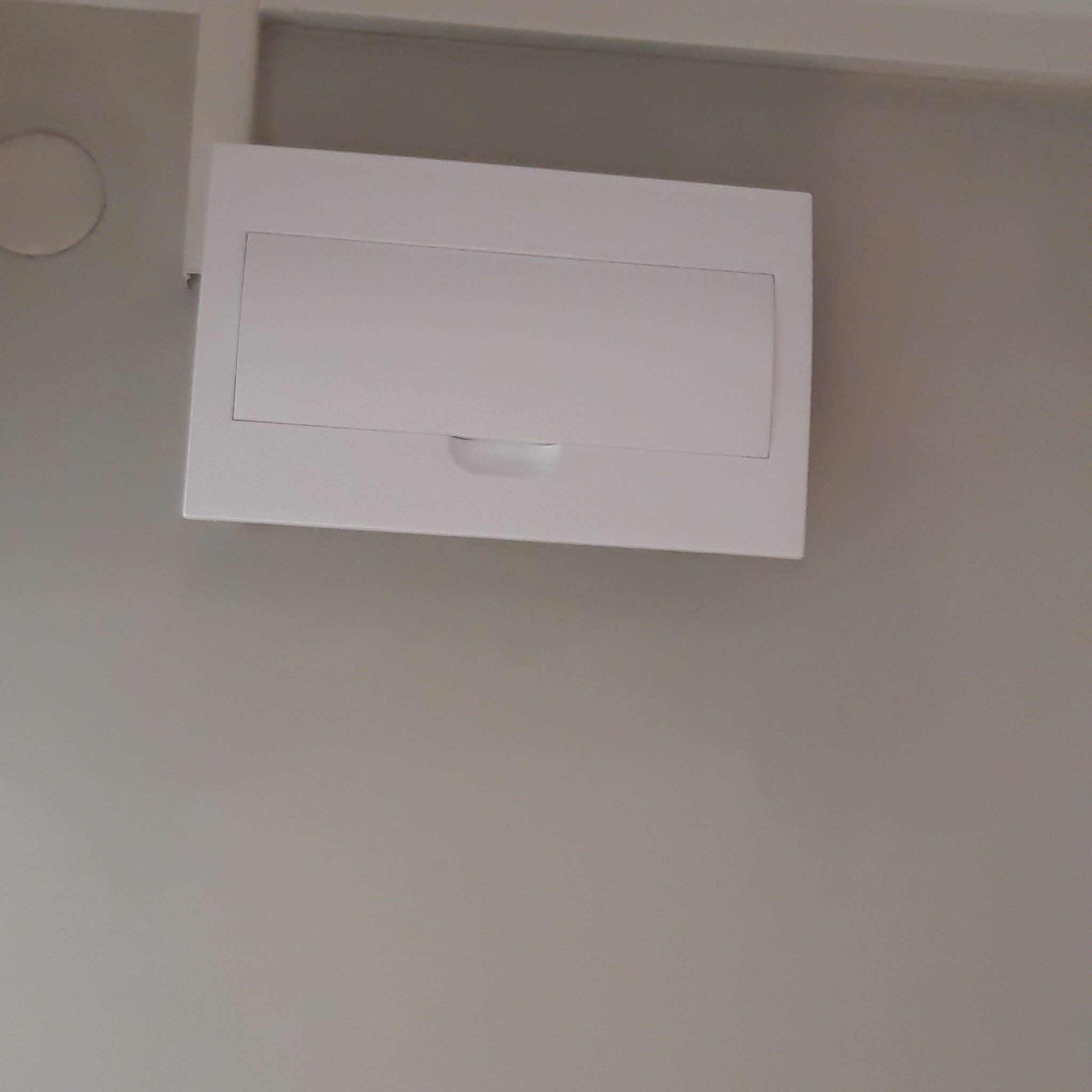 Biała skrzynka elektryczna natynkowa z klapką, zamontowana na szarej ścianie, z widocznym fragmentem listwy przypodłogowej.