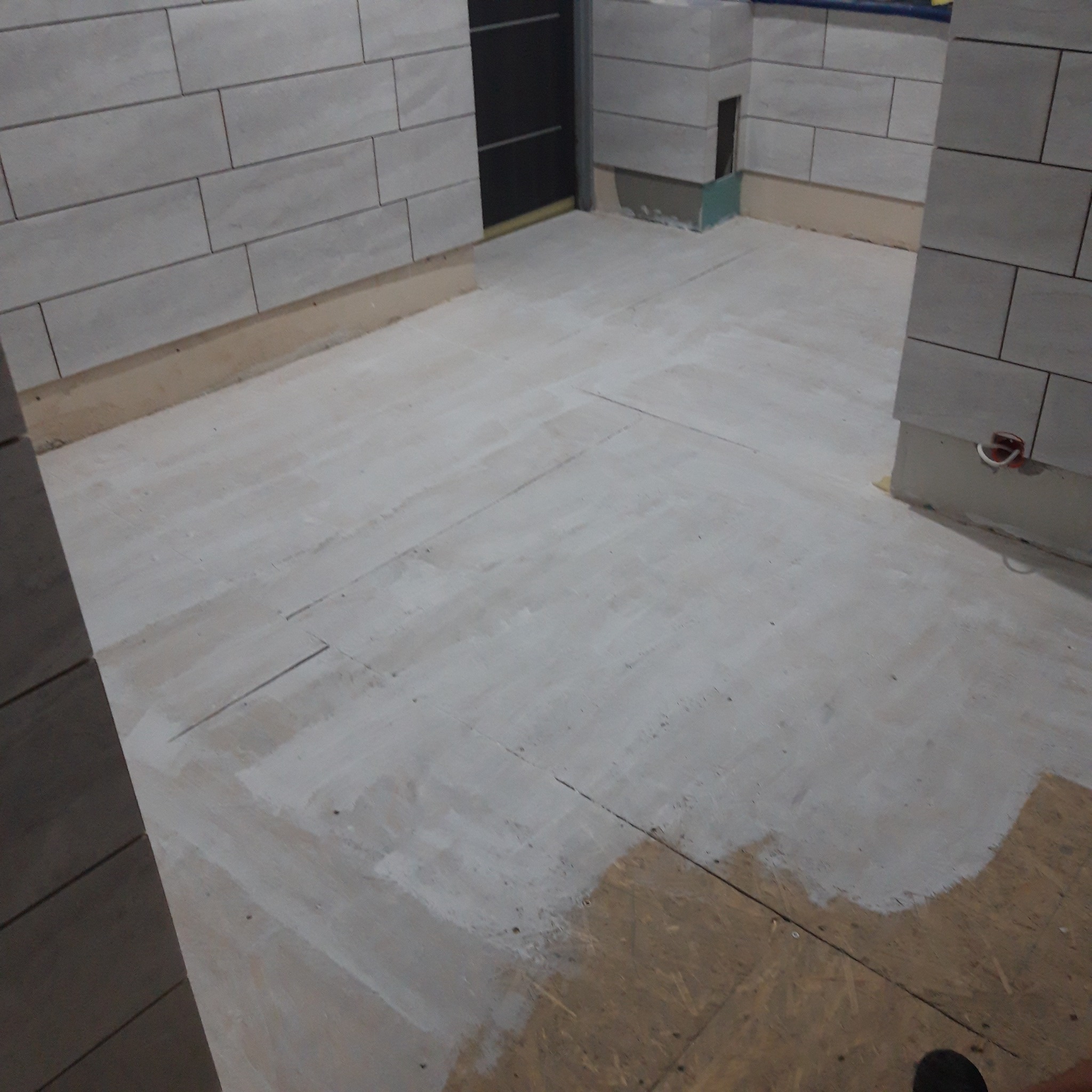 Surowy stan podłogi z płyt OSB po pierwszym malowaniu na biało, ściany wyłożone szarą płytką imitującą cegłę, widoczne kable elektryczne i fragmenty instalacji.