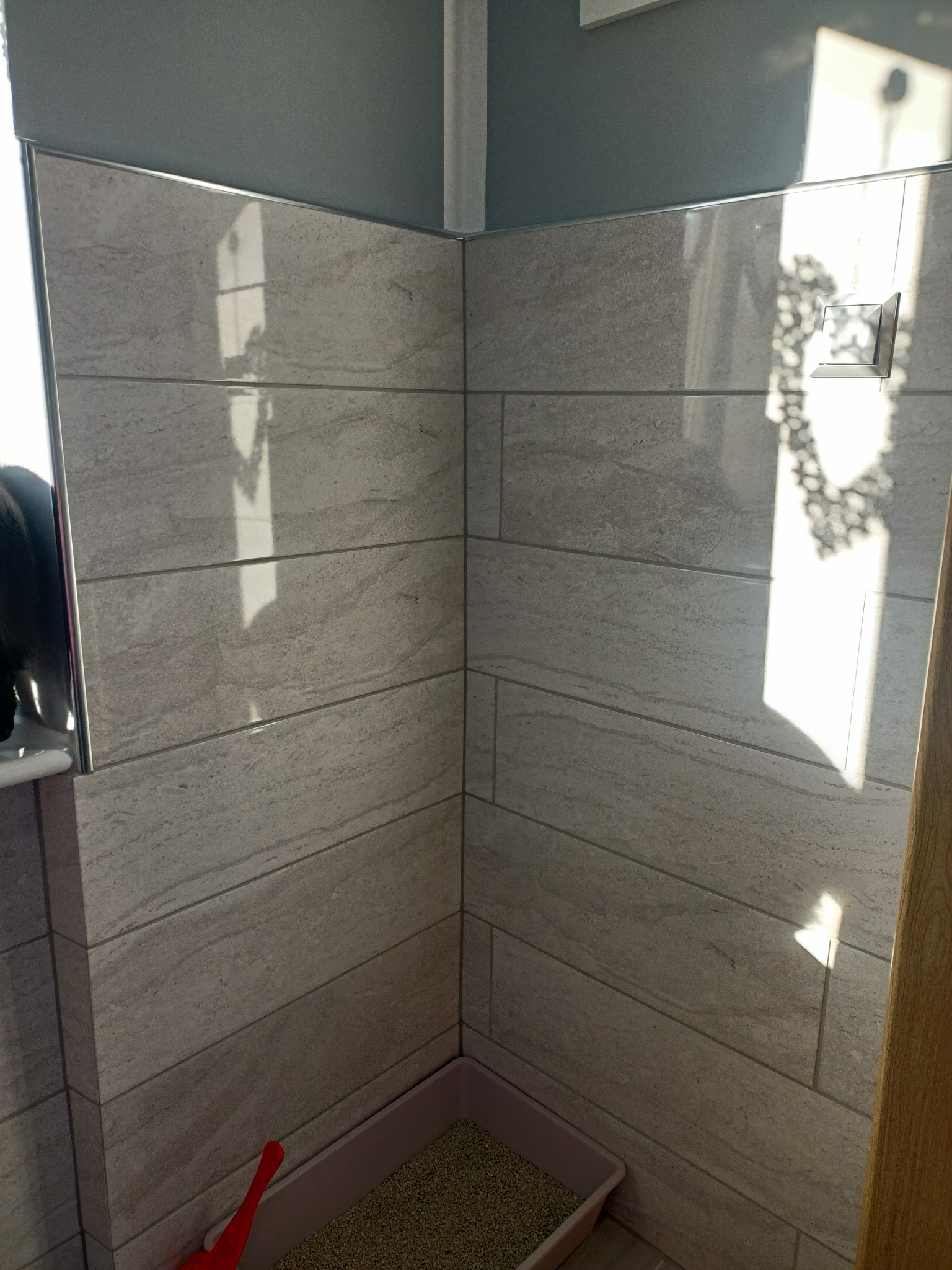 Wykończona łazienka z szarymi płytkami imitującymi kamień, widoczny fragment lustra, toalety i kuweta dla kota w rogu.