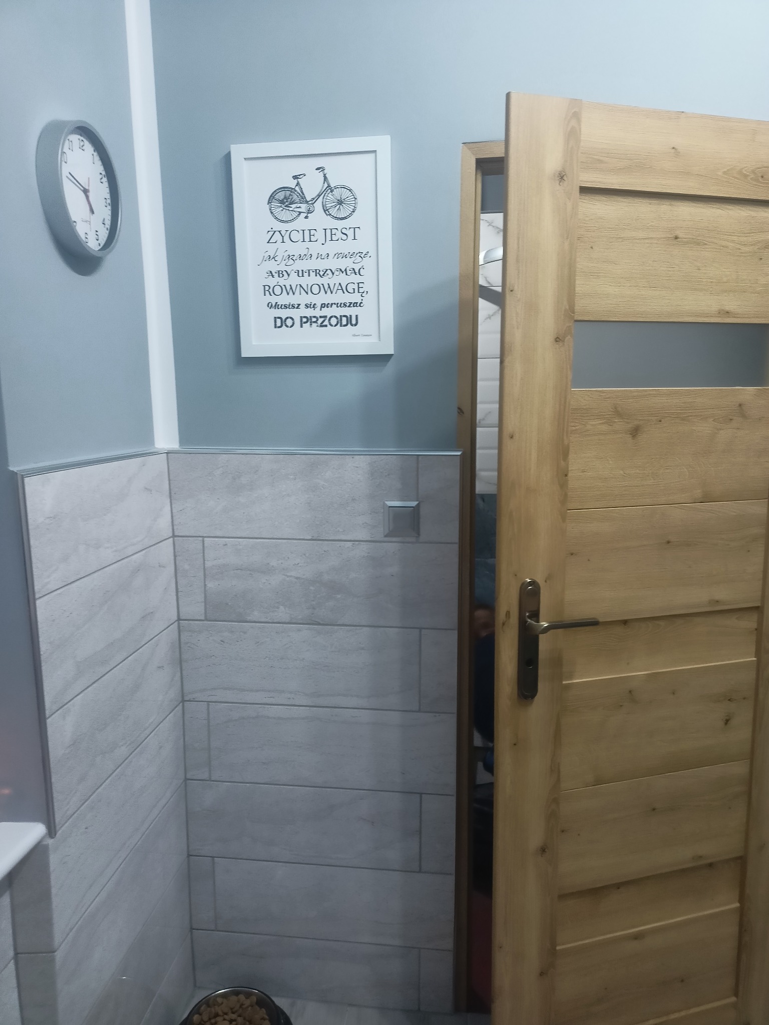 Wykończona łazienka z szarymi płytkami imitującymi drewno, drewniane drzwi z przeszkleniem, zegar na ścianie oraz obrazek z rowerem i sentencją.