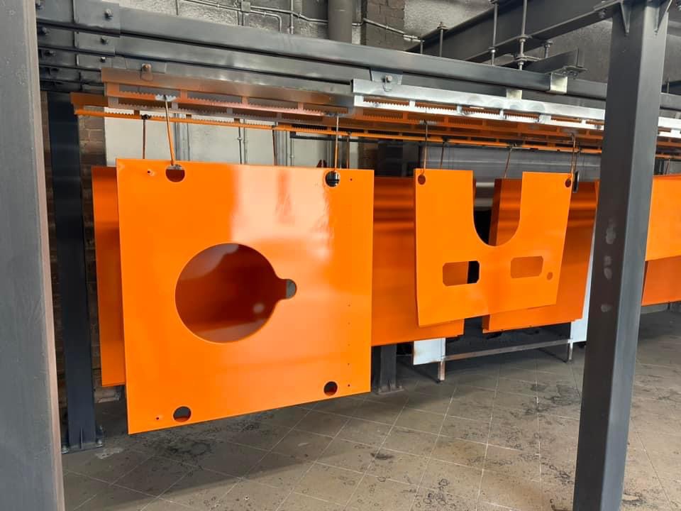 Pomarańczowe, metalowe elementy po lakierowaniu proszkowym, zawieszone na linii produkcyjnej.