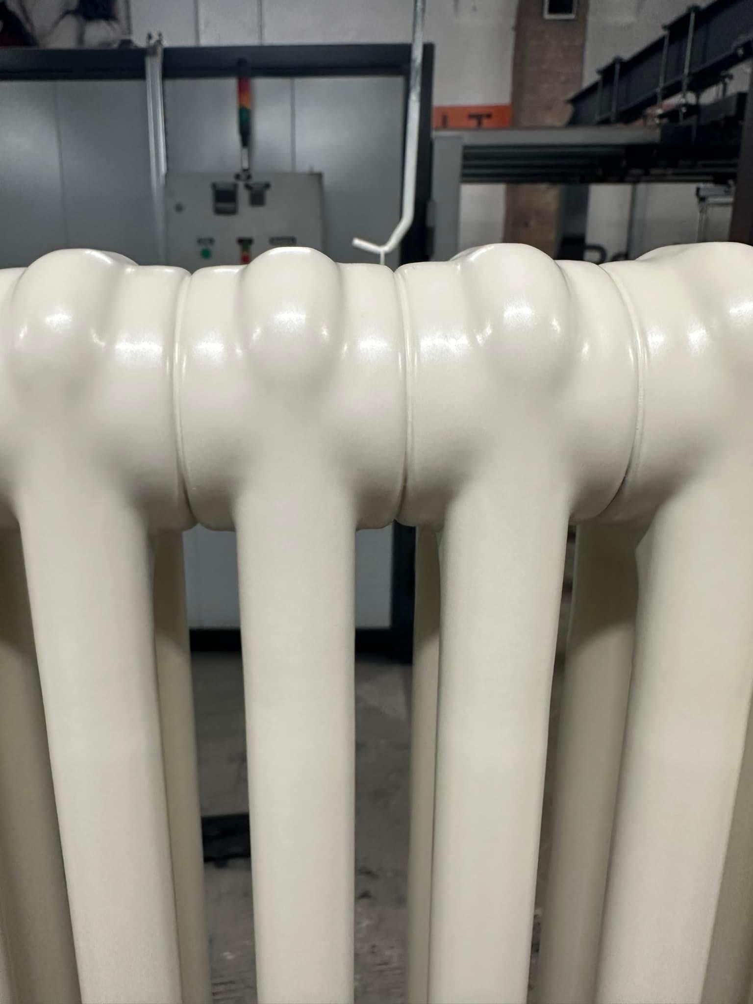 Biały, żeliwny grzejnik po renowacji, z widocznymi śladami świeżego lakieru, na tle hali produkcyjnej.