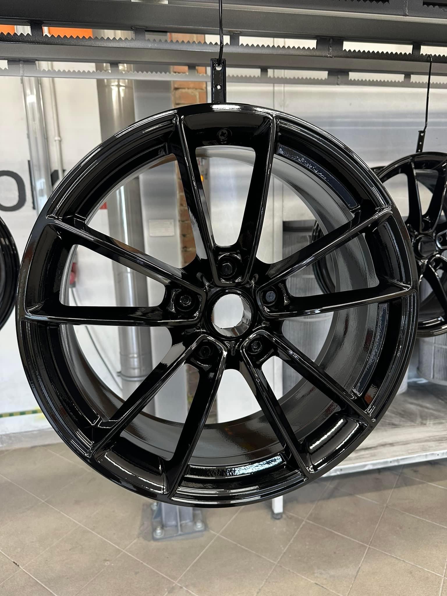 Błyszcząca, czarna felga aluminiowa zawieszona na lakierni proszkowej, z widocznymi refleksami światła na gładkiej powierzchni.