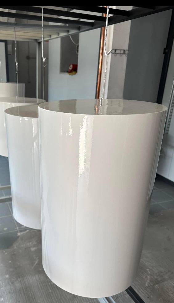 Białe, cylindryczne elementy zawieszone na hakach po lakierowaniu, widoczny połysk świeżej powłoki w oświetlonym pomieszczeniu.