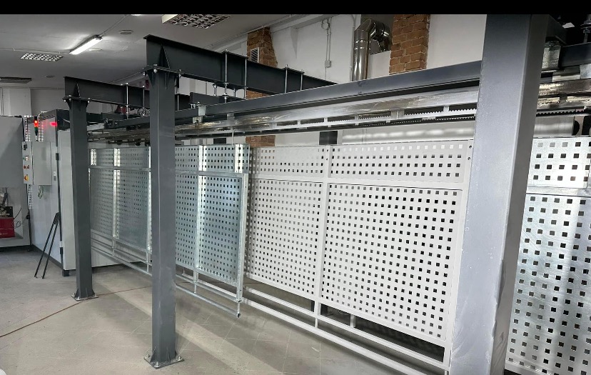 Automatyczna linia lakiernicza z zawieszonymi elementami z perforowanej blachy, widoczne sterowanie elektryczne i elementy konstrukcyjne hali.