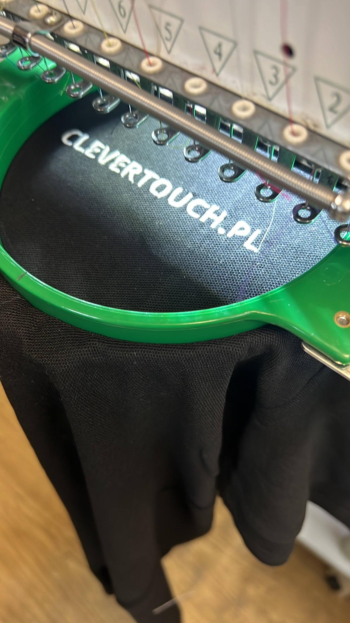 Proces haftowania napisu 'CLEVERTOUCH.PL' na czarnej tkaninie w Krakowie, z użyciem profesjonalnej maszyny hafciarskiej z zielonym tamborkiem.