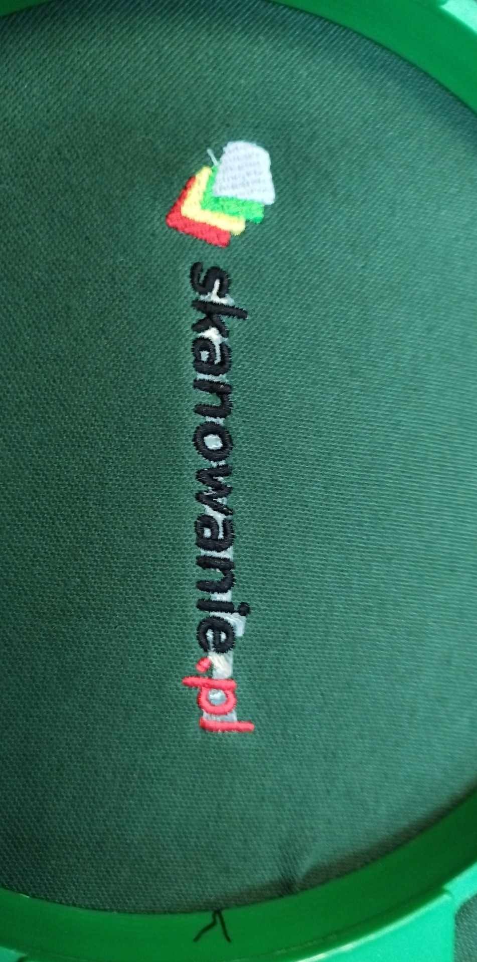 Zielona tkanina z wyhaftowanym kolorowym logo i adresem strony 'skanowanienieruchomosci.pl' w ramce do haftowania. Detal haftu.