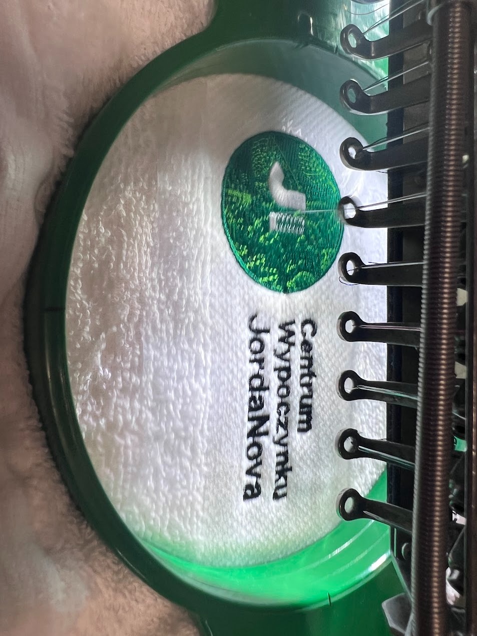 Haftowany ręcznik z logo 'Centrum Wypoczynku JordaNova' w zielonej ramce maszyny hafciarskiej. Widoczne igły i mechanizm maszyny. Detal haftu.