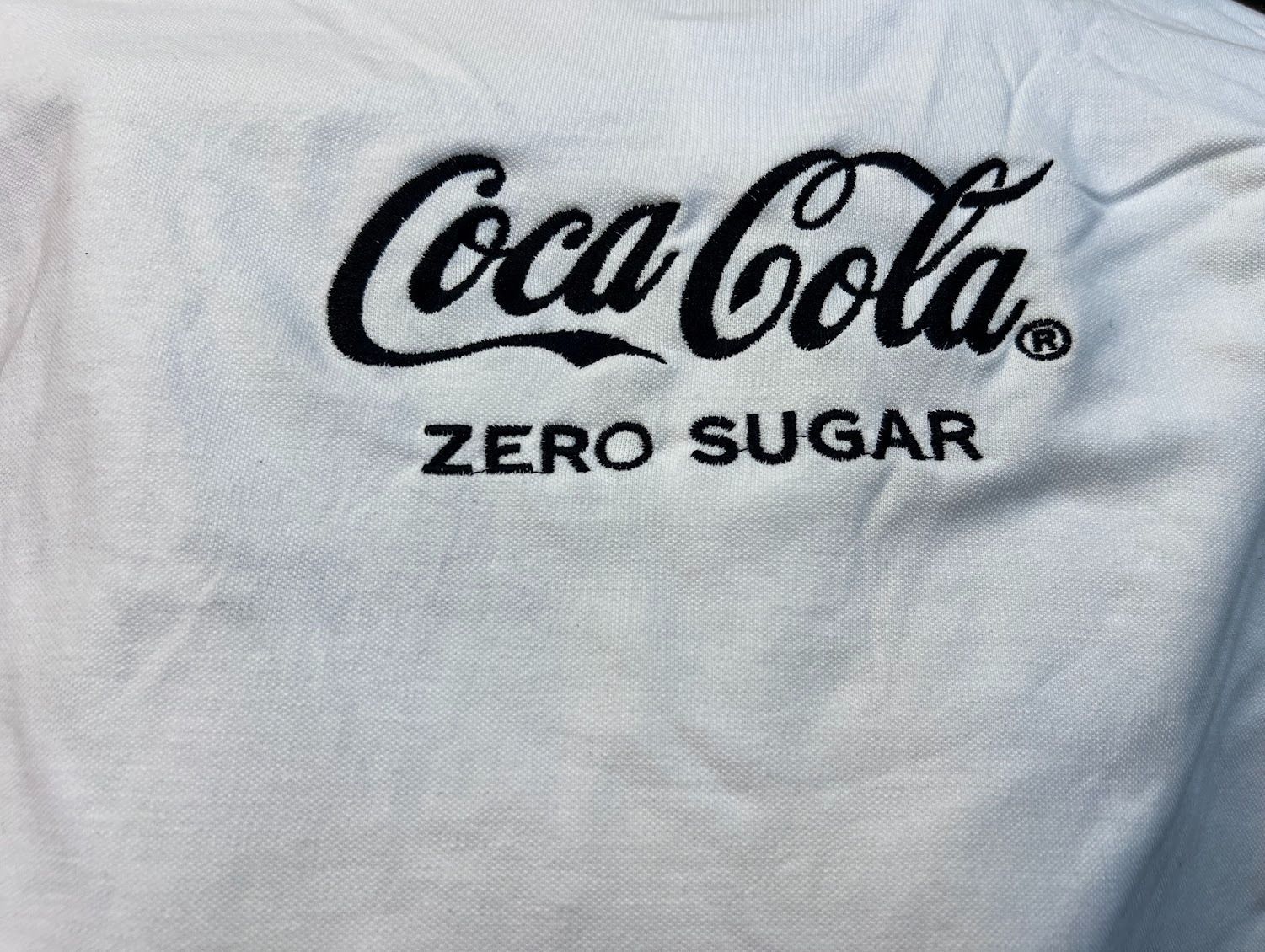 Biała koszulka polo z czarnym haftem 'Coca-Cola ZERO SUGAR', prezentująca detale wykonania i jakość materiału.