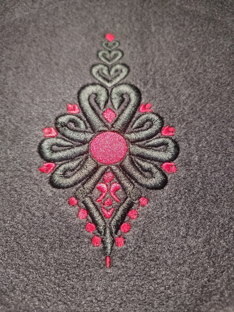 Szczegółowe ujęcie ciemnoszarej tkaniny z wyhaftowanym, symetrycznym wzorem w odcieniach czerni i różu. Wzór przypomina stylizowane kwiaty i serca.