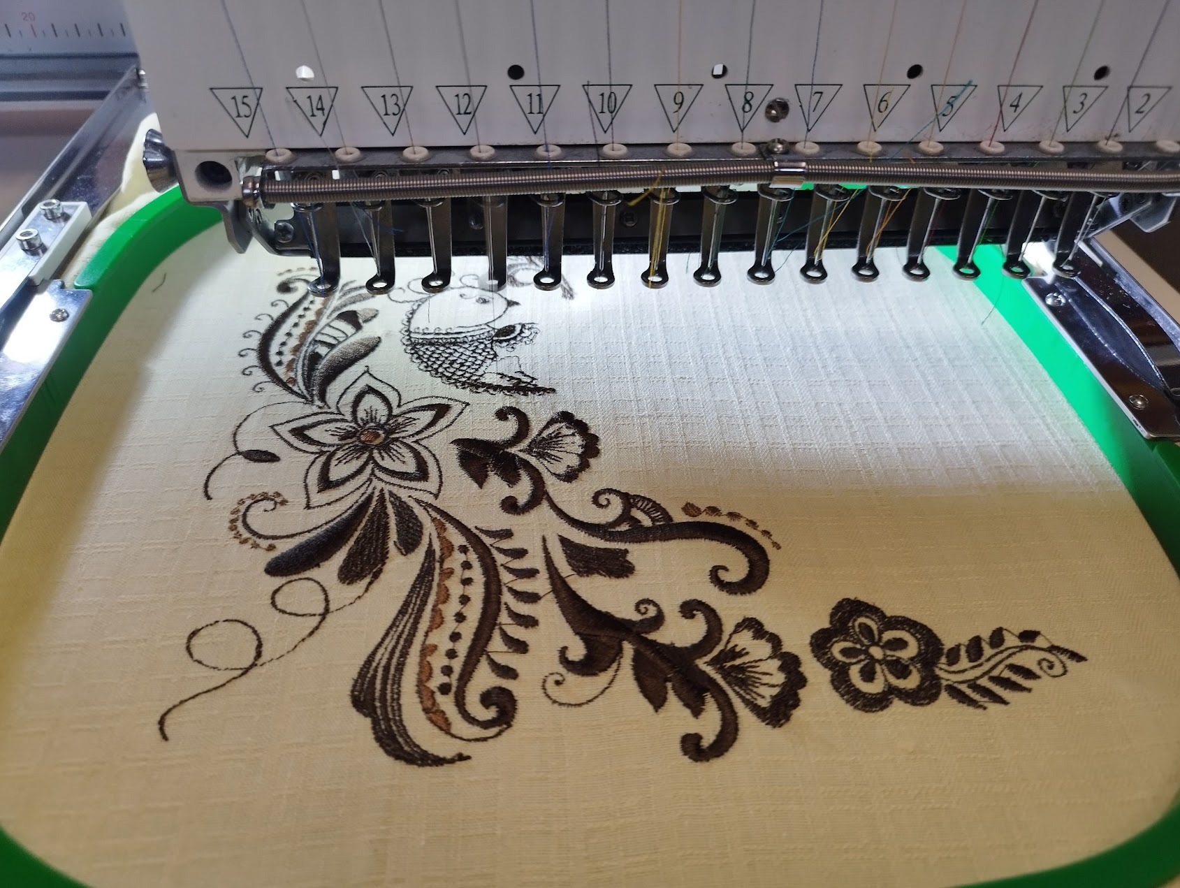 Haftowany wzór kwiatowy z motywem ryby na beżowej tkaninie, widoczny fragment maszyny hafciarskiej z igłami i zieloną ramką. Detal haftu w zbliżeniu.