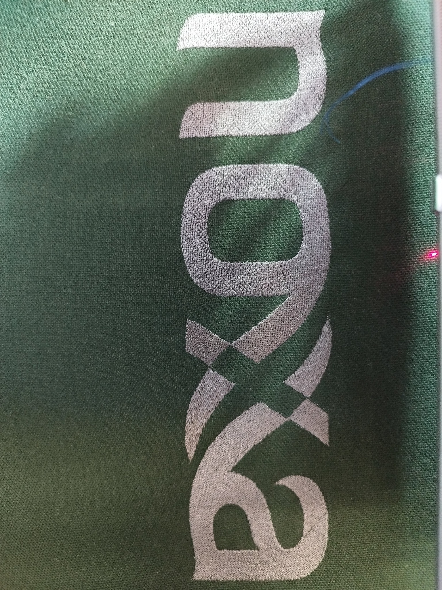 Zielona tkanina z wyhaftowanym srebrną nicią pionowym napisem 'axon'. Widoczna faktura materiału i precyzja wykonania haftu.