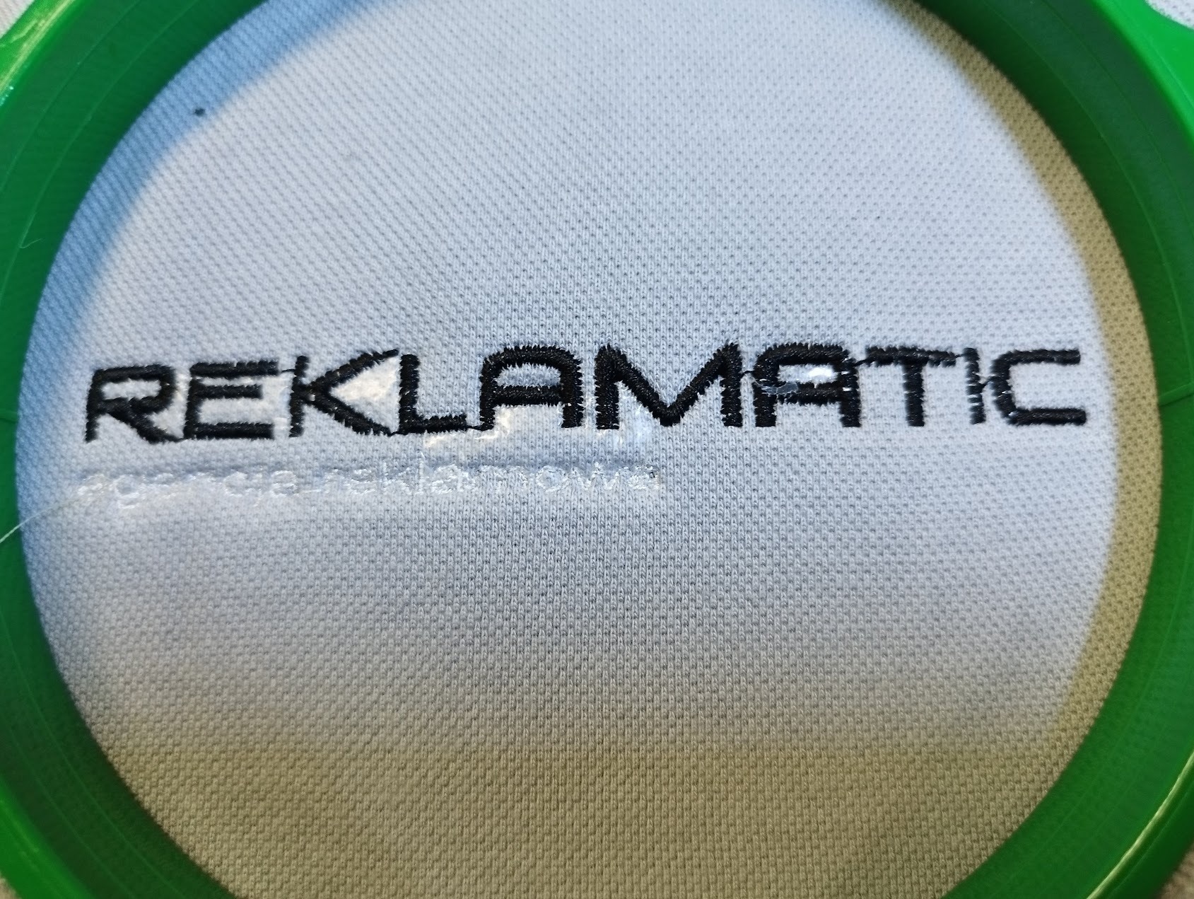 Haftowany napis 'REKLAMATIC' na białym materiale w zielonej ramce. Widoczna struktura tkaniny i detale haftu. Bliskie ujęcie, ostre światło.