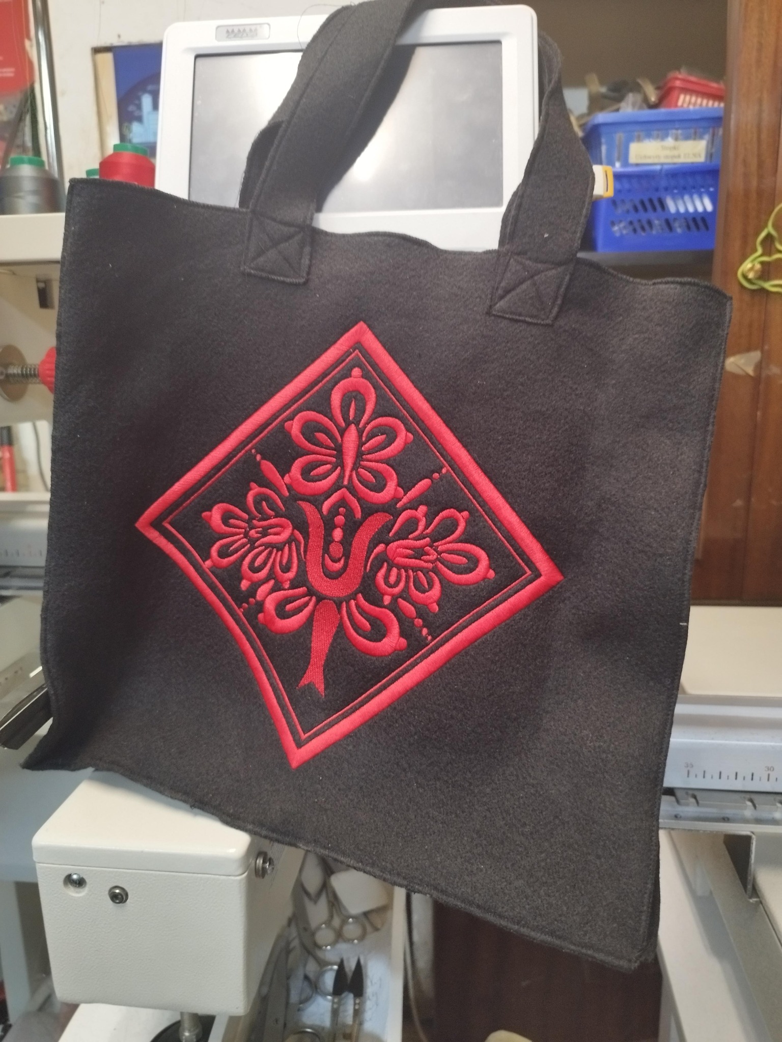 Czarna torba z filcu z czerwonym, kwadratowym haftem o motywie roślinnym, leżąca na stole obok maszyny do haftowania. Widoczne nici i akcesoria.