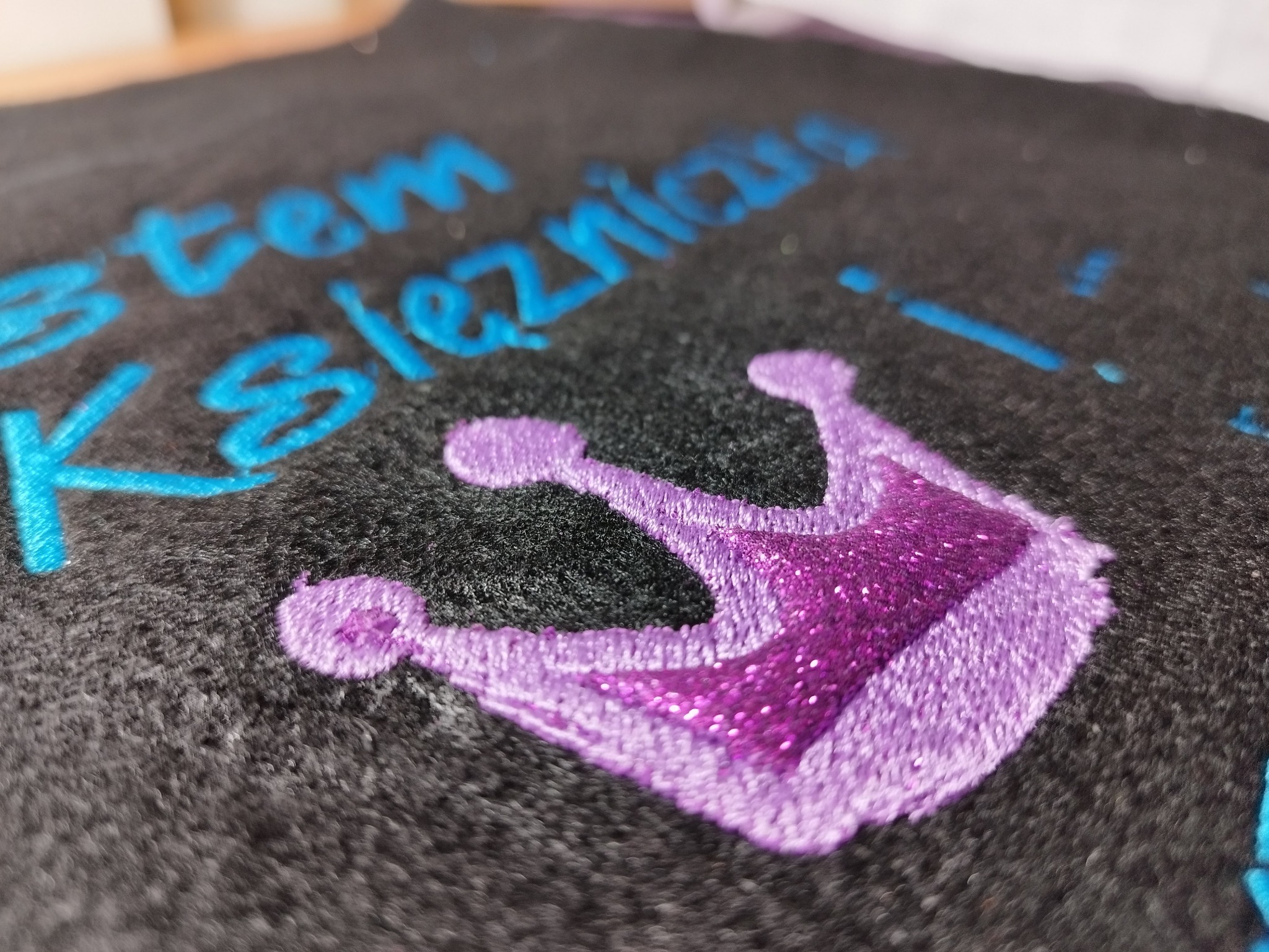 Czarny materiał z wyhaftowanym, błyszczącym, fioletowym motywem korony i niebieskim napisem 'Jestem Księżniczką'. Detal haftu, bliskie ujęcie.