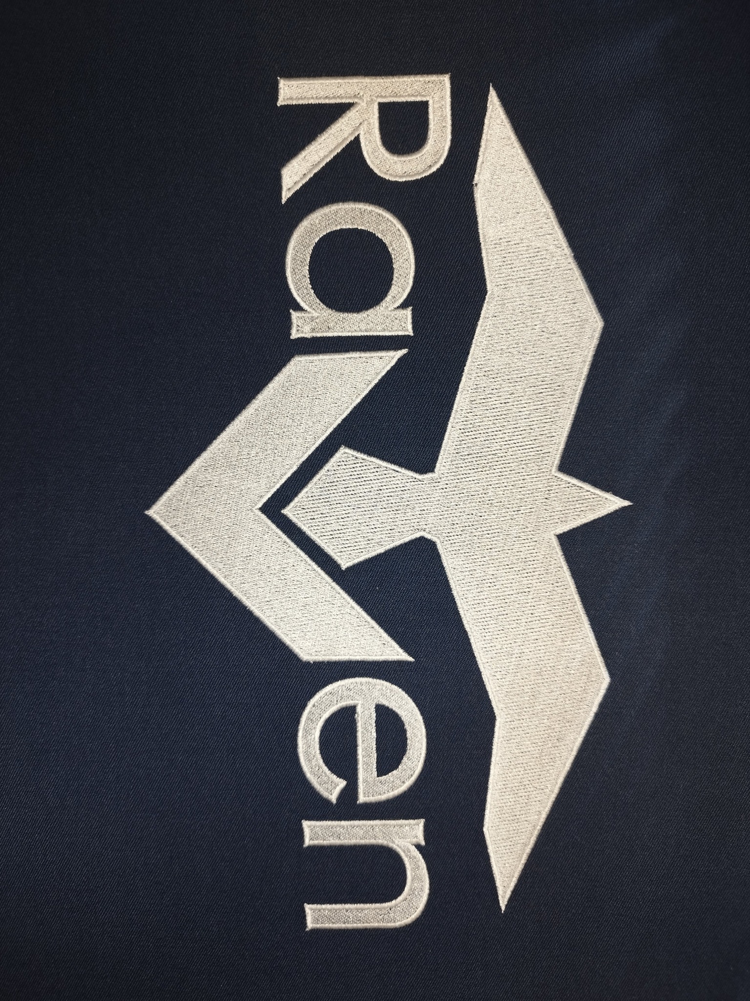 Ciemnoniebieska tkanina z wyhaftowanym pionowo białym napisem 'Raven' i symbolem ptaka. Detal haftu, widoczna faktura materiału i nici.