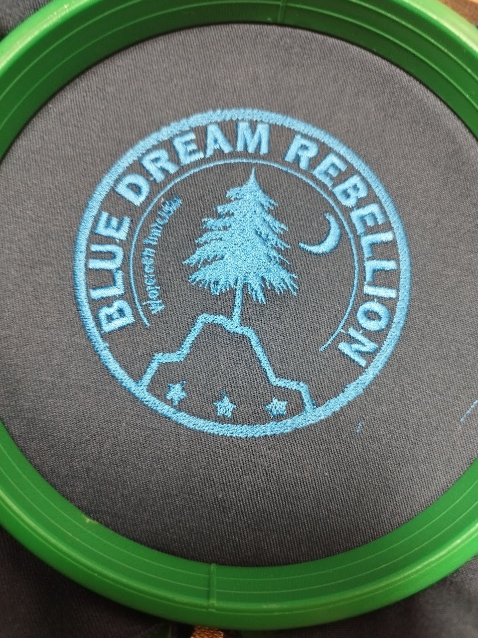 Niebieski haft z motywem drzewa i napisem 'BLUE DREAM REBELLION' na szarej tkaninie, umieszczony w zielonej tamborku. Detalowe wykończenie, bliski kadr.