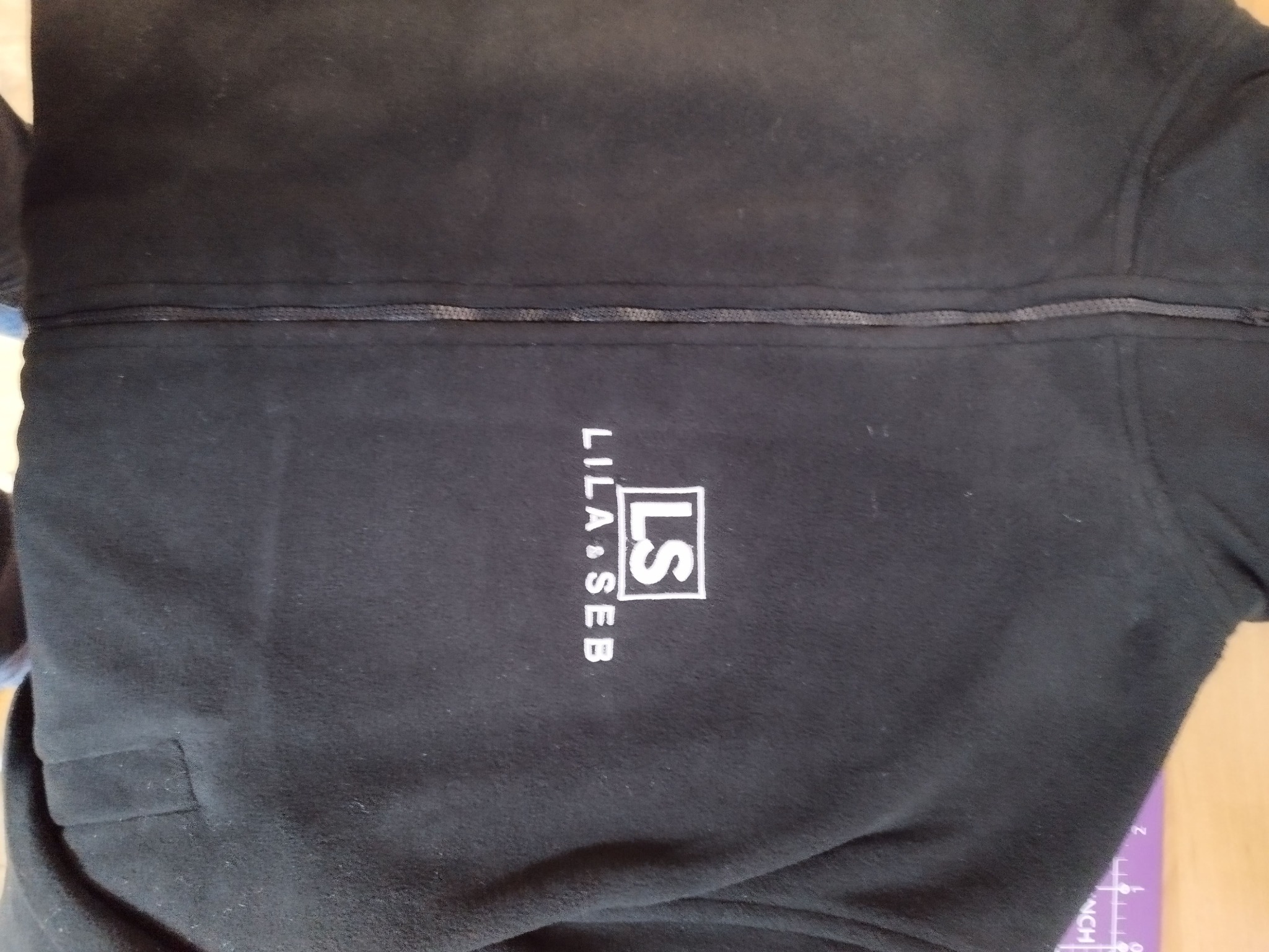 Czarna bluza polarowa z haftowanym logo 'LS LILA & SEB' na piersi, widoczny zamek błyskawiczny i kieszeń. Ujęcie z bliska, lekko z góry.