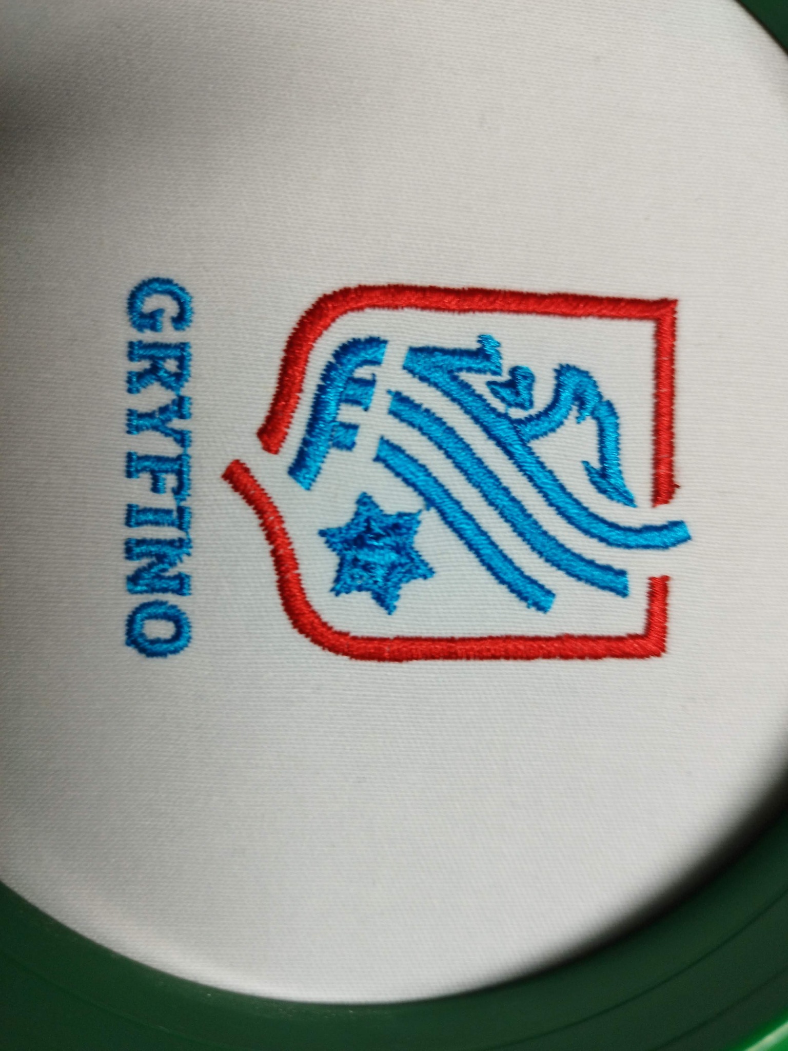 Haftowany, kolorowy herb 'GRYFINO' na białym materiale, umieszczony w zielonej ramce. Detaliczne wykonanie, widoczne ściegi. Herb w kolorach czerwonym i niebieskim.