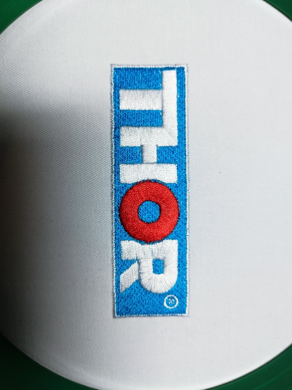 Detal haftu z napisem 'THOR' na białym materiale. Napis w kolorach: biały, czerwony i niebieski. Widoczna faktura haftu i precyzja wykonania.