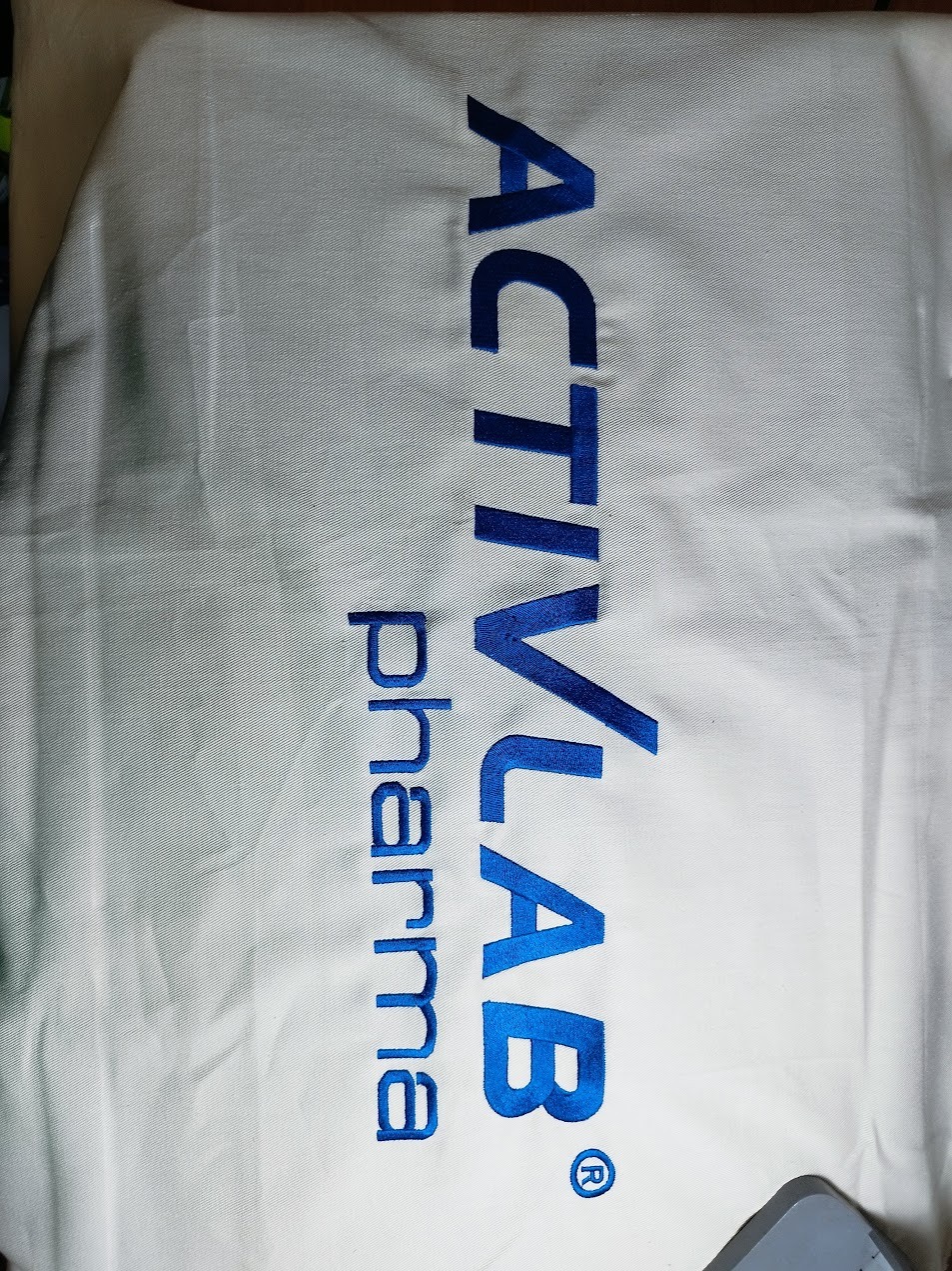 Biała tkanina z wyhaftowanym, pionowym napisem 'ACTIVLAB pharma' w kolorze niebieskim. Widoczne detale ściegów i faktura materiału.