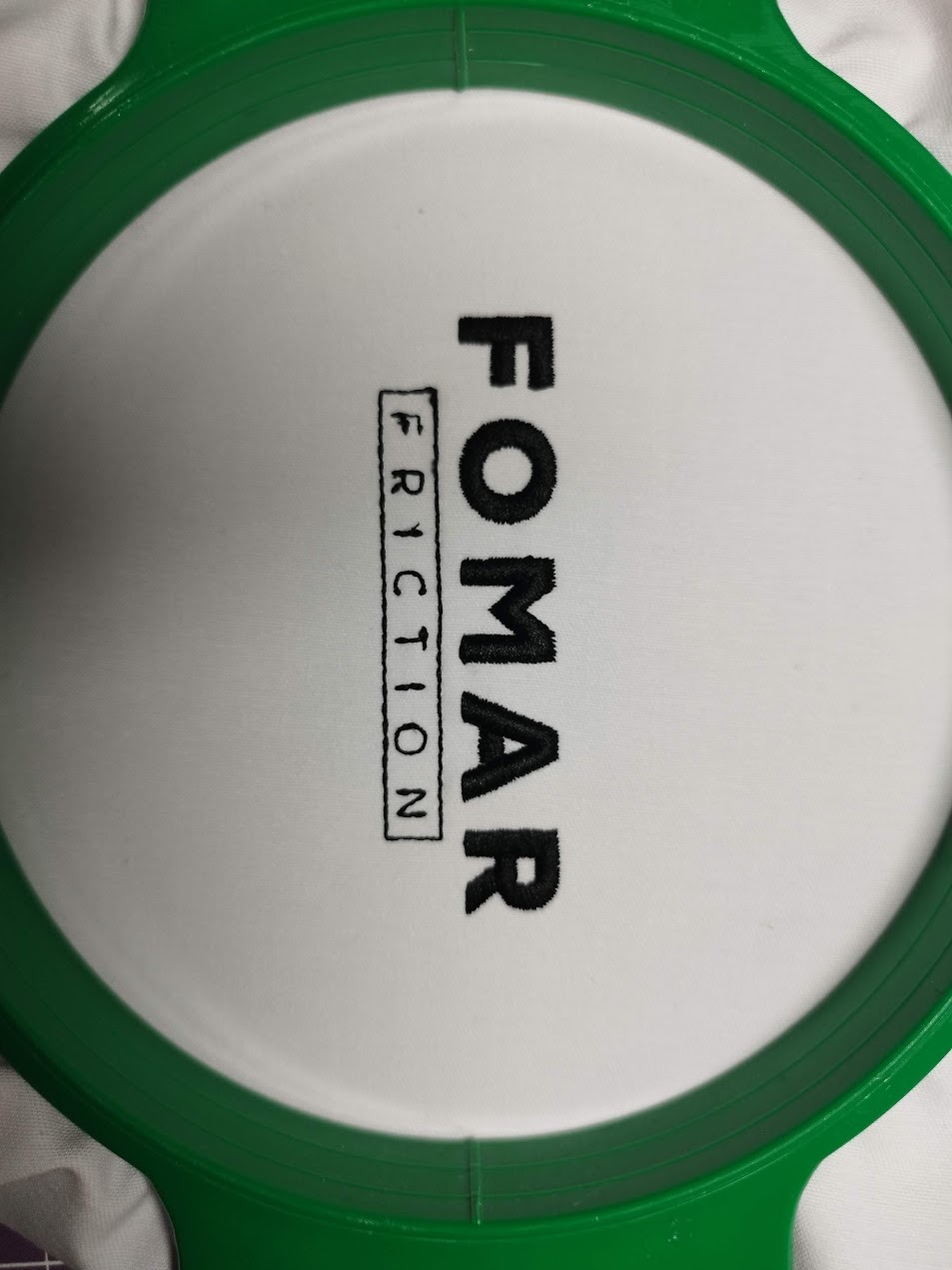 Haftowany napis 'FOMAR FRICTION' czarną nicią na białym materiale, umieszczony w zielonej tamborku. Widok z góry, detale ściegów.