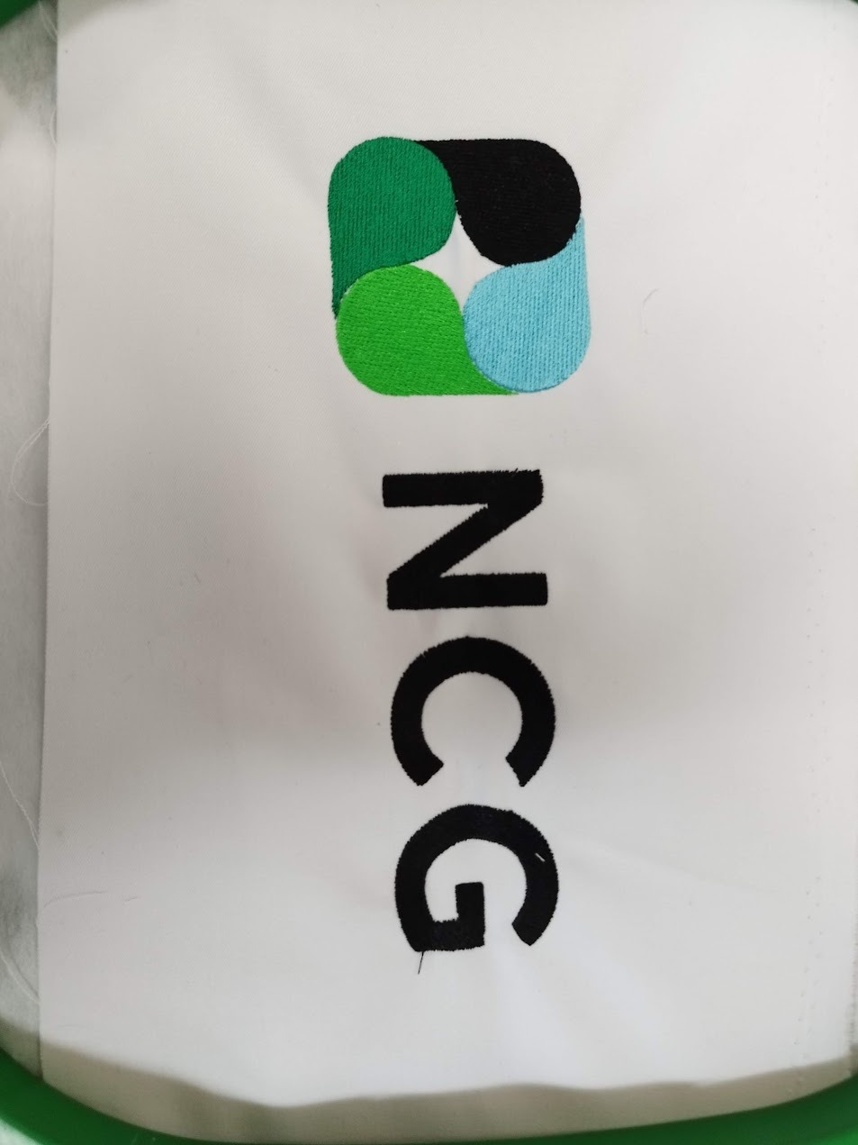 Wyhaftowane logo NCG na białym materiale, widoczne detale ściegów i kolorów: zielony, czarny i błękitny. Całość umieszczona w zielonej ramce.