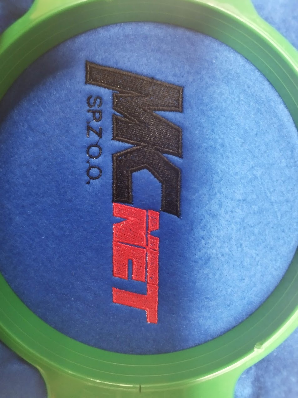 Niebieski materiał w tamborku z wyhaftowanym czarno-czerwonym logo 'MK NET SP. Z O.O.'. Detal haftu maszynowego z bliska, prezentacja gotowego produktu.