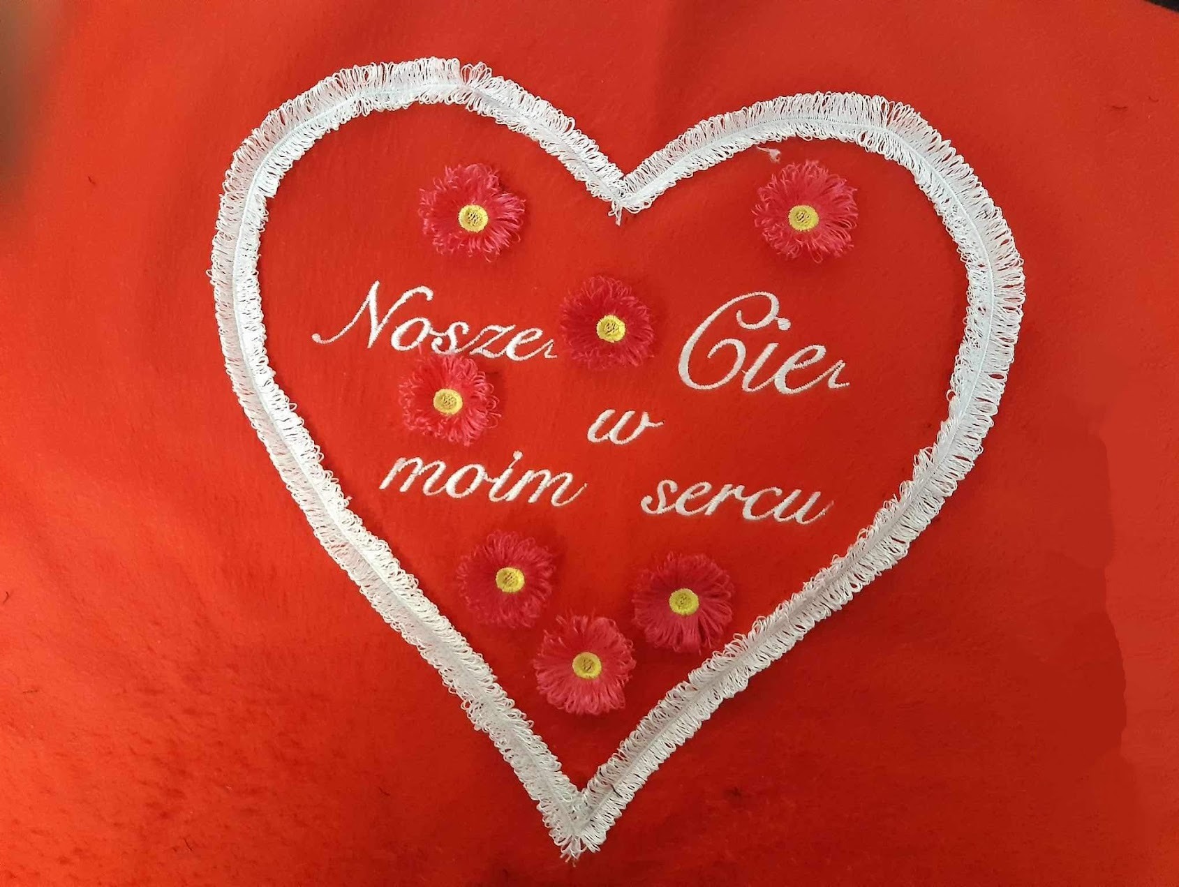 Czerwona poduszka z haftowanym białym sercem, kwiatami i napisem 'Noszę Cię w moim sercu'. Dekoracyjny, ręcznie wykonany prezent walentynkowy.