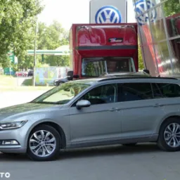 Szary Volkswagen Passat Variant zaparkowany przed salonem samochodowym z czerwonym samochodem ciężarowym w tle, widoczne logo VW na budynku.