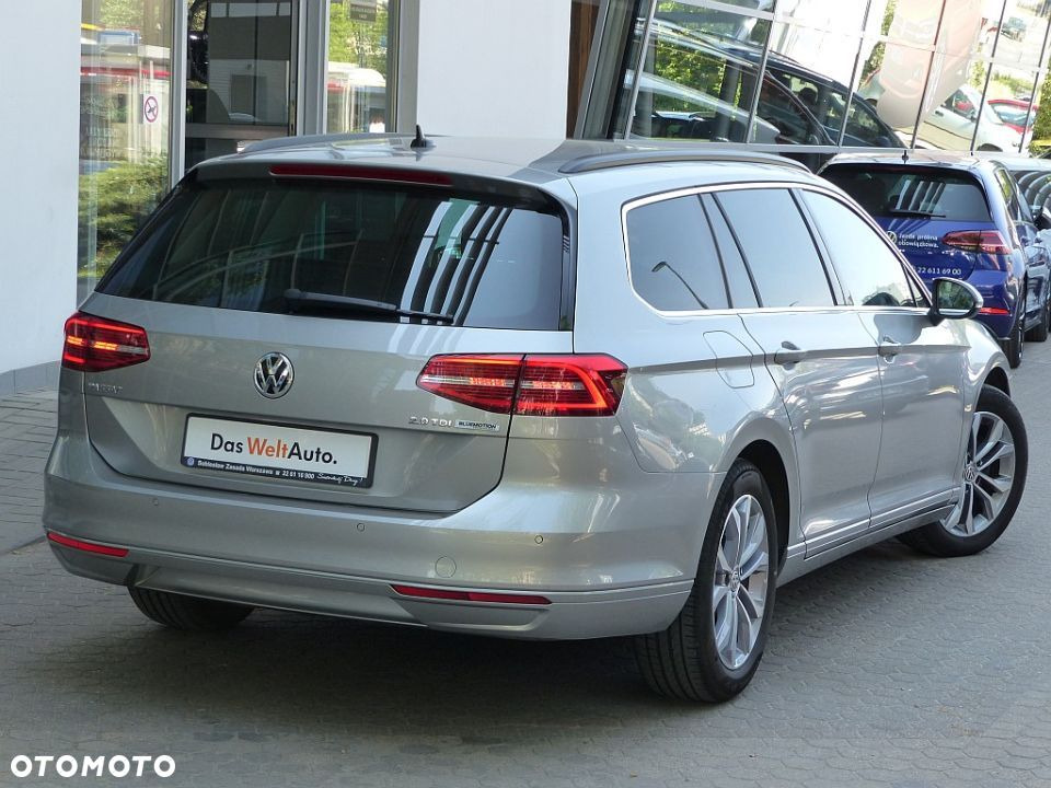 Srebrny samochód kombi Volkswagen Passat Variant z widocznym logo Das WeltAuto, zaparkowany przed salonem samochodowym z odbiciami w szklanej fasadzie.