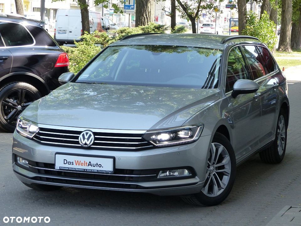 Jasnoszary Volkswagen Passat Variant z salonu Das WeltAuto, zaparkowany na chodniku, z widocznym logo VW na grillu i czarnym SUV-em w tle.