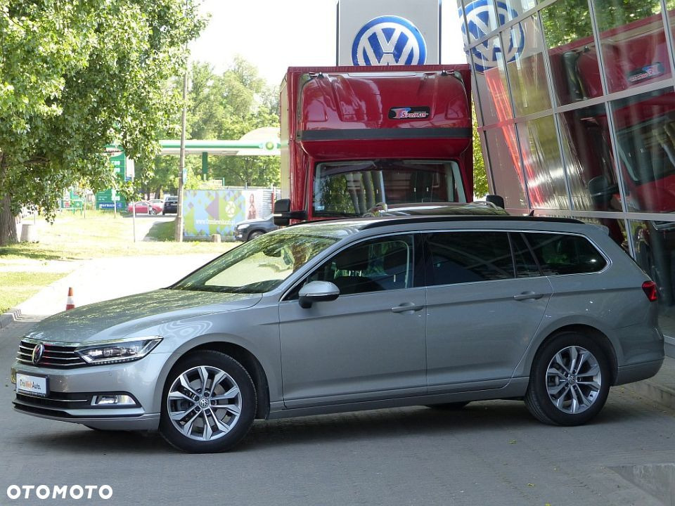 Szary Volkswagen Passat Variant zaparkowany przed salonem samochodowym z czerwonym samochodem ciężarowym w tle, widoczne logo VW na budynku.