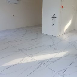 Robert Siara - Jasne, marmurowe płytki podłogowe w nowym, pustym pomieszczeniu z oknem i klimatyzacją. Widoczne przygotowane punkty elektryczne i hydrauliczne na ścianach.