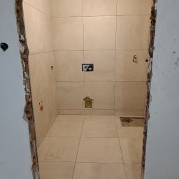 Firma Remont-Budowlana RS-BUD - Wnętrze łazienki w trakcie prac wykończeniowych: ułożone jasne płytki na ścianach i podłodze, widoczne instalacje, surowy stan ścian przy wejściu, oświetlenie sufitowe.