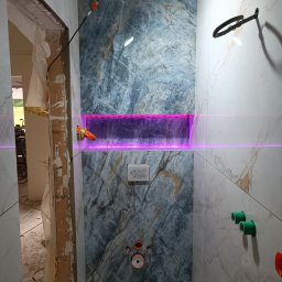 Firma Remont-Budowlana RS-BUD - Wnętrze łazienki w trakcie prac wykończeniowych. Widoczne płytki imitujące marmur, podświetlana wnęka, instalacja wodna i elektryczna. Ościeżnica drzwiowa w stanie surowym.