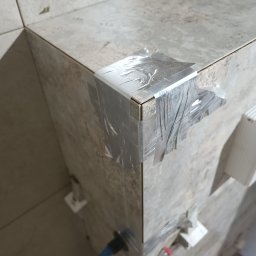 Firma Remont-Budowlana RS-BUD - Fragment narożnika obłożonego płytkami, zabezpieczonego srebrną taśmą. Widoczne krzyżyki dystansowe i fragment narzędzia glazurniczego. Ujęcie z bliska, z góry.