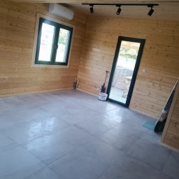 Firma Remont-Budowlana RS-BUD - Wnętrze wykończonego pomieszczenia z drewnianymi ścianami, szarą podłogą z płytek, oknem i drzwiami. Widoczne oświetlenie punktowe i klimatyzacja.