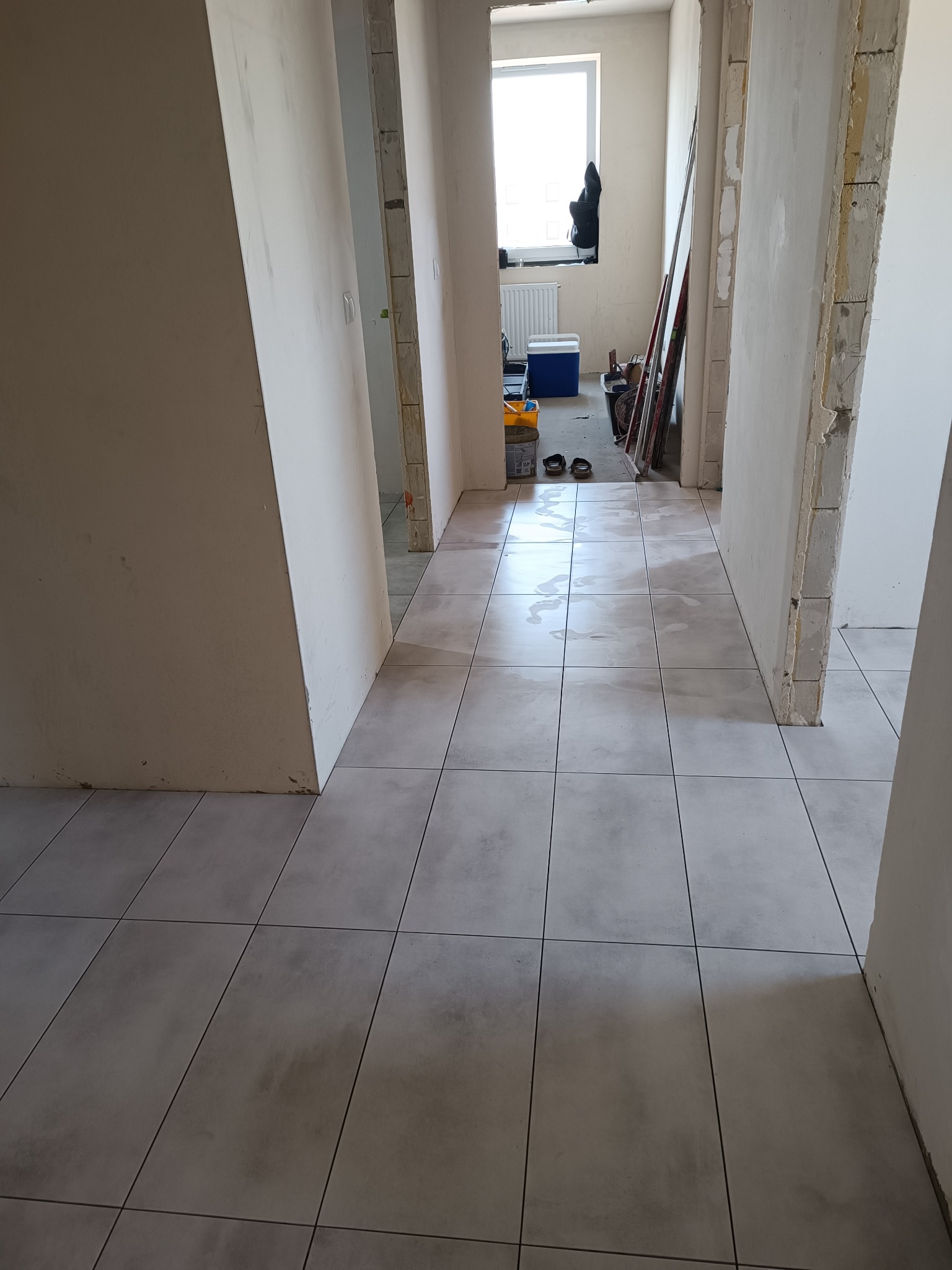 Ułożone, szare płytki podłogowe w korytarzu w trakcie remontu. Widoczne surowe ściany i przygotowane otwory drzwiowe. W tle okno z widokiem na zewnątrz.