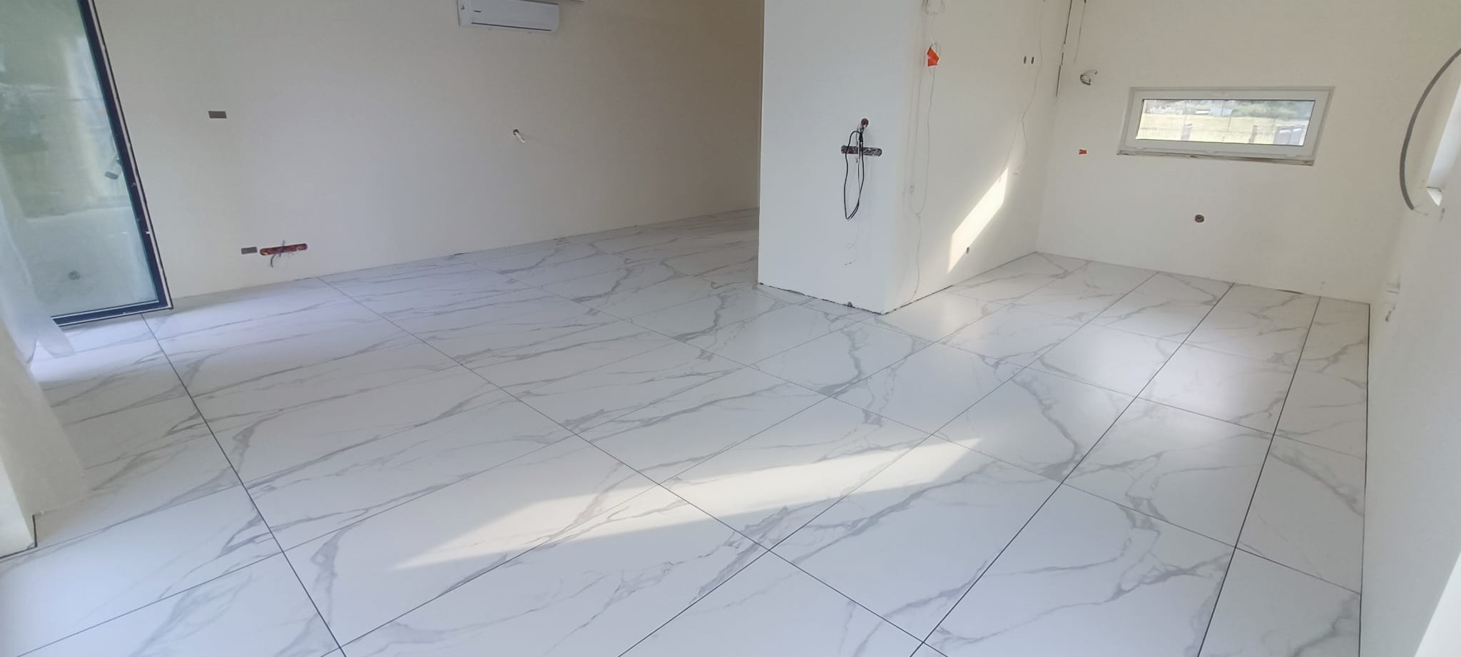 Jasne, marmurowe płytki podłogowe w nowym, pustym pomieszczeniu z oknem i klimatyzacją. Widoczne przygotowane punkty elektryczne i hydrauliczne na ścianach.