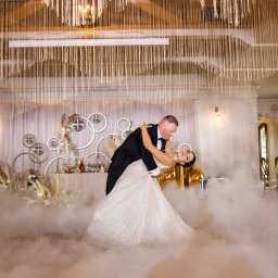 PHU KAR-EVENTS - Para młoda tańczy pierwszy taniec w chmurach dymu na sali weselnej z dekoracjami w stylu glamour, na tle stołu z dekoracjami i złotymi elementami.