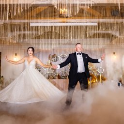 PHU KAR-EVENTS - Para młoda w trakcie pierwszego tańca na parkiecie spowitym sztuczną mgłą, dekoracje sali weselnej w tle.