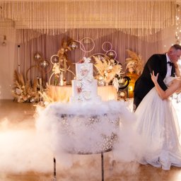 PHU KAR-EVENTS - Para młoda tańczy w otoczeniu iskier i sztucznej mgły, na pierwszym planie tort weselny z kwiatowymi dekoracjami, w tle dekoracje z suszonych traw i okrągłych elementów.