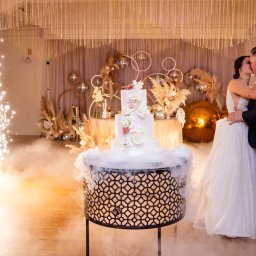 PHU KAR-EVENTS - Para młoda całuje się na parkiecie podczas wesela, w tle tort weselny na dekoracyjnym postumencie, efekty specjalne w postaci iskier i dymu, fotograf robiący zdjęcia.