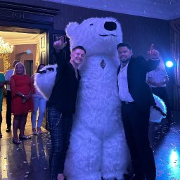 PHU KAR-EVENTS - Dwóch mężczyzn w eleganckich strojach pozuje z dużym, białym pluszowym niedźwiedziem w sali balowej z żyrandolem i dekoracjami świetlnymi, konfetti na podłodze.