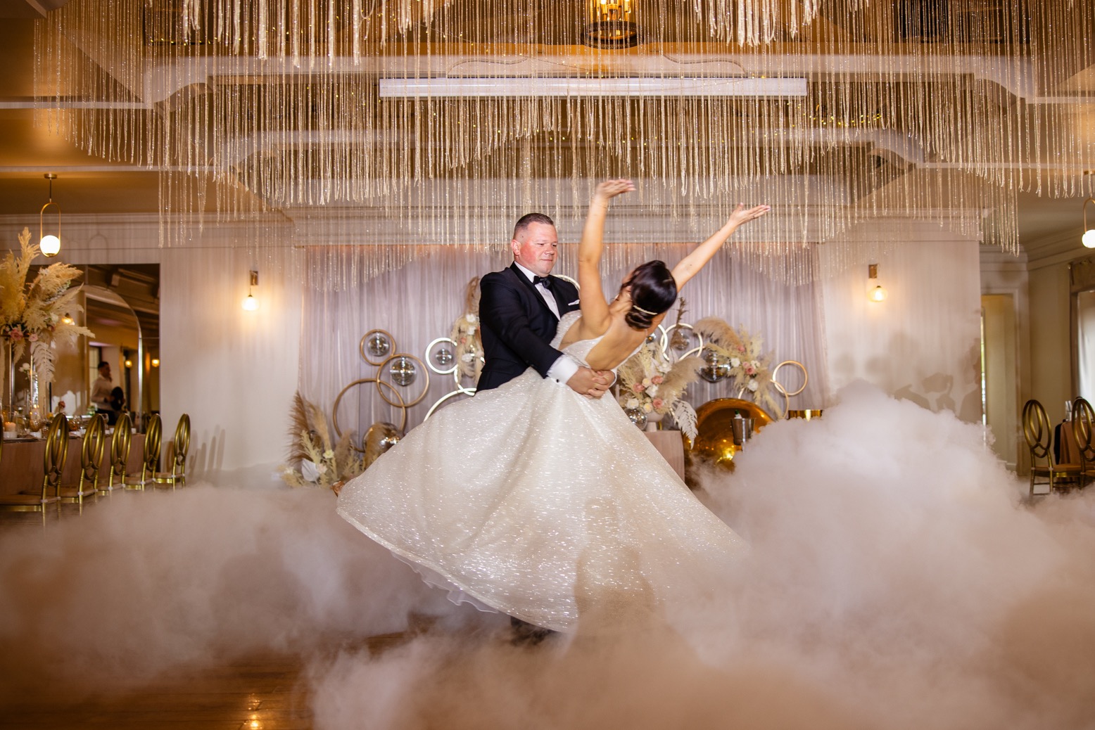 Para młoda w tańcu w chmurze dymu na sali weselnej z dekoracjami w stylu boho i kryształowym żyrandolem.