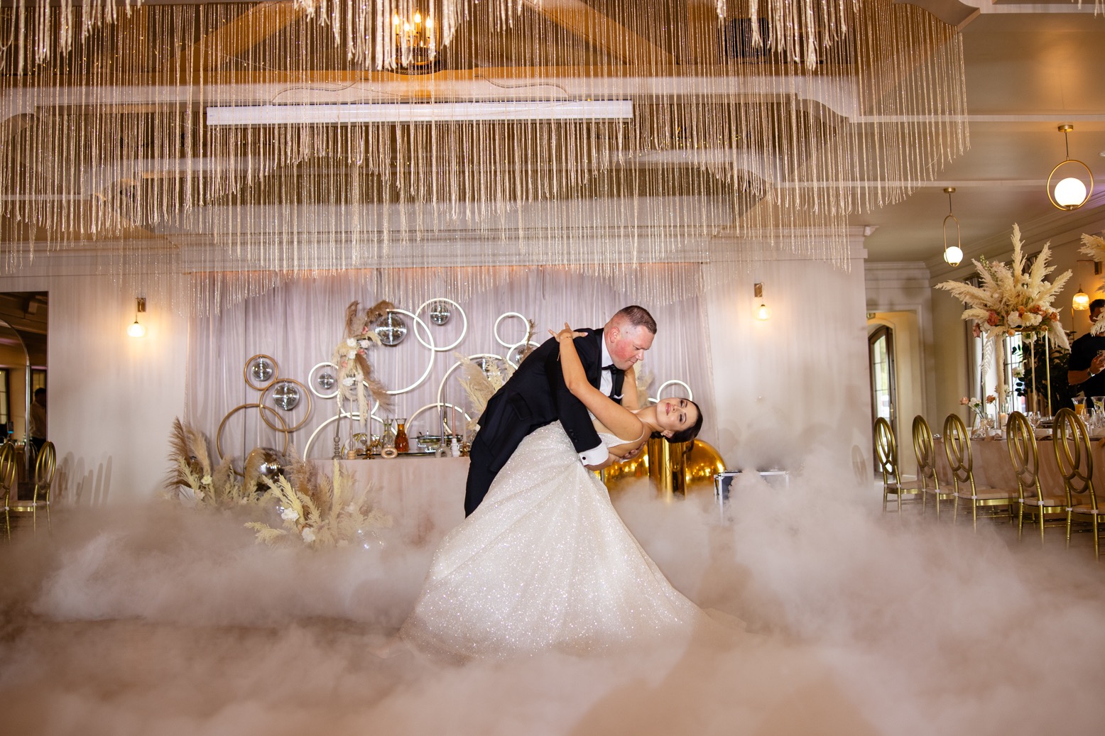 Para młoda tańczy pierwszy taniec w chmurach dymu na sali weselnej z dekoracjami w stylu glamour, na tle stołu z dekoracjami i złotymi elementami.