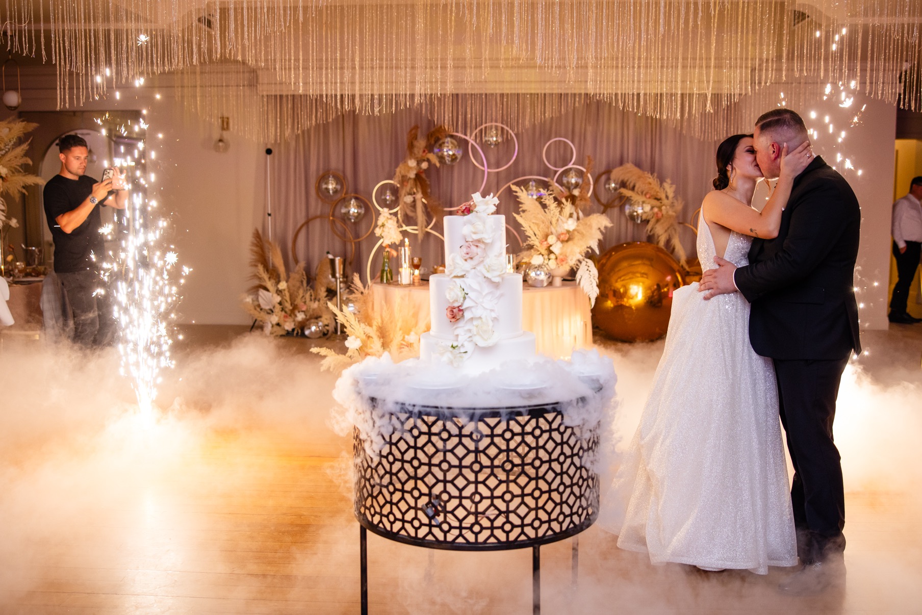 Para młoda całuje się na parkiecie podczas wesela, w tle tort weselny na dekoracyjnym postumencie, efekty specjalne w postaci iskier i dymu, fotograf robiący zdjęcia.