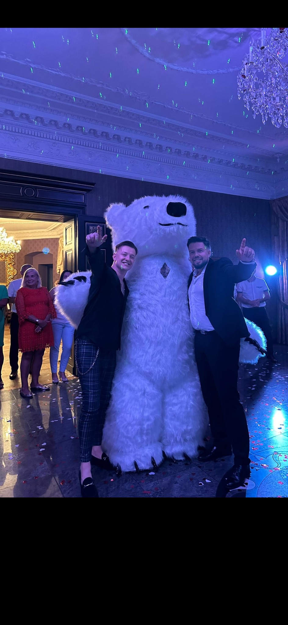 Dwóch mężczyzn w eleganckich strojach pozuje z dużym, białym pluszowym niedźwiedziem w sali balowej z żyrandolem i dekoracjami świetlnymi, konfetti na podłodze.