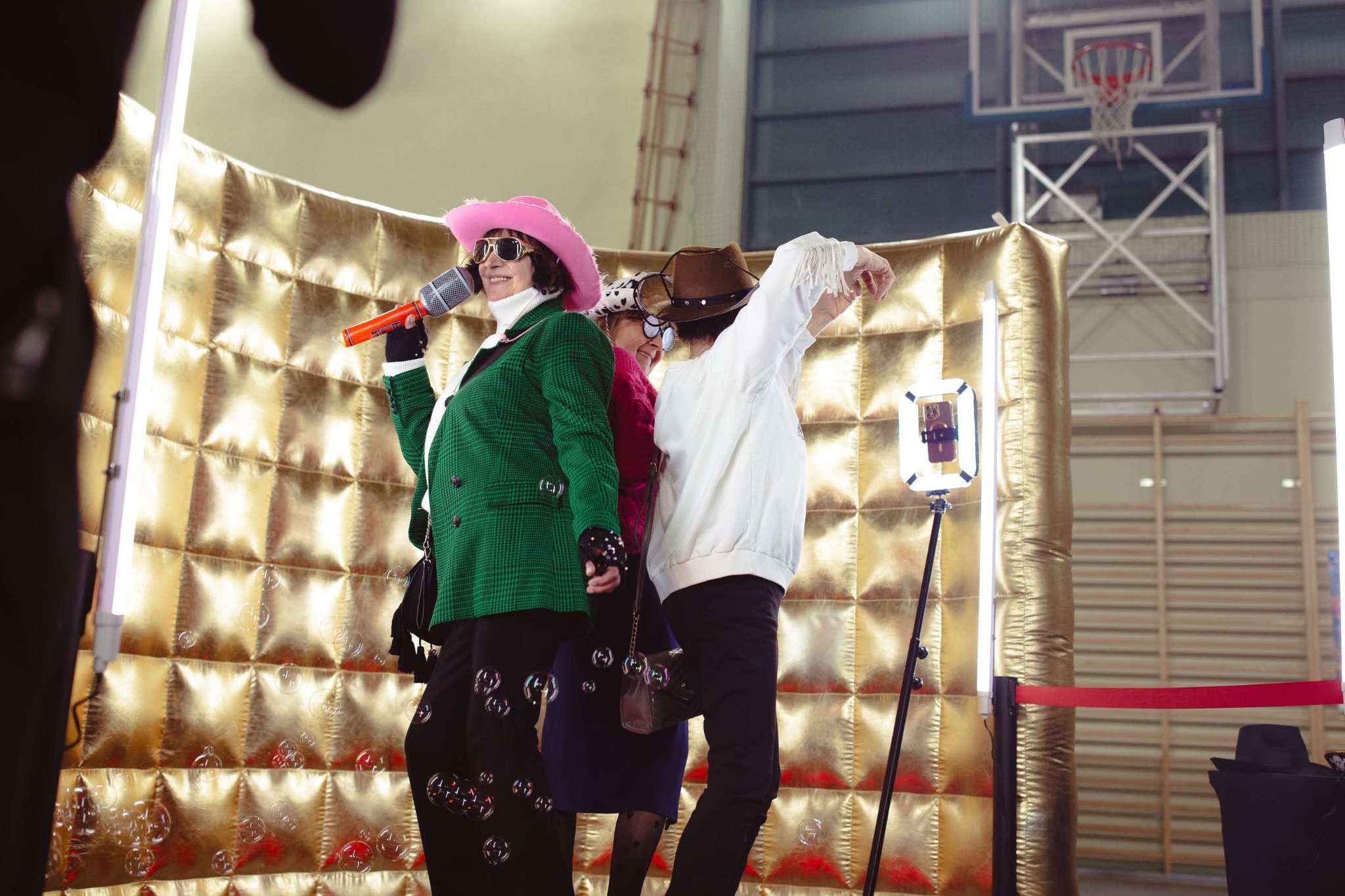 Trzy osoby w przebraniach pozują wewnątrz złotej fotobudki z mikrofonem, oświetleniem LED i bańkami mydlanymi, na tle sali gimnastycznej.